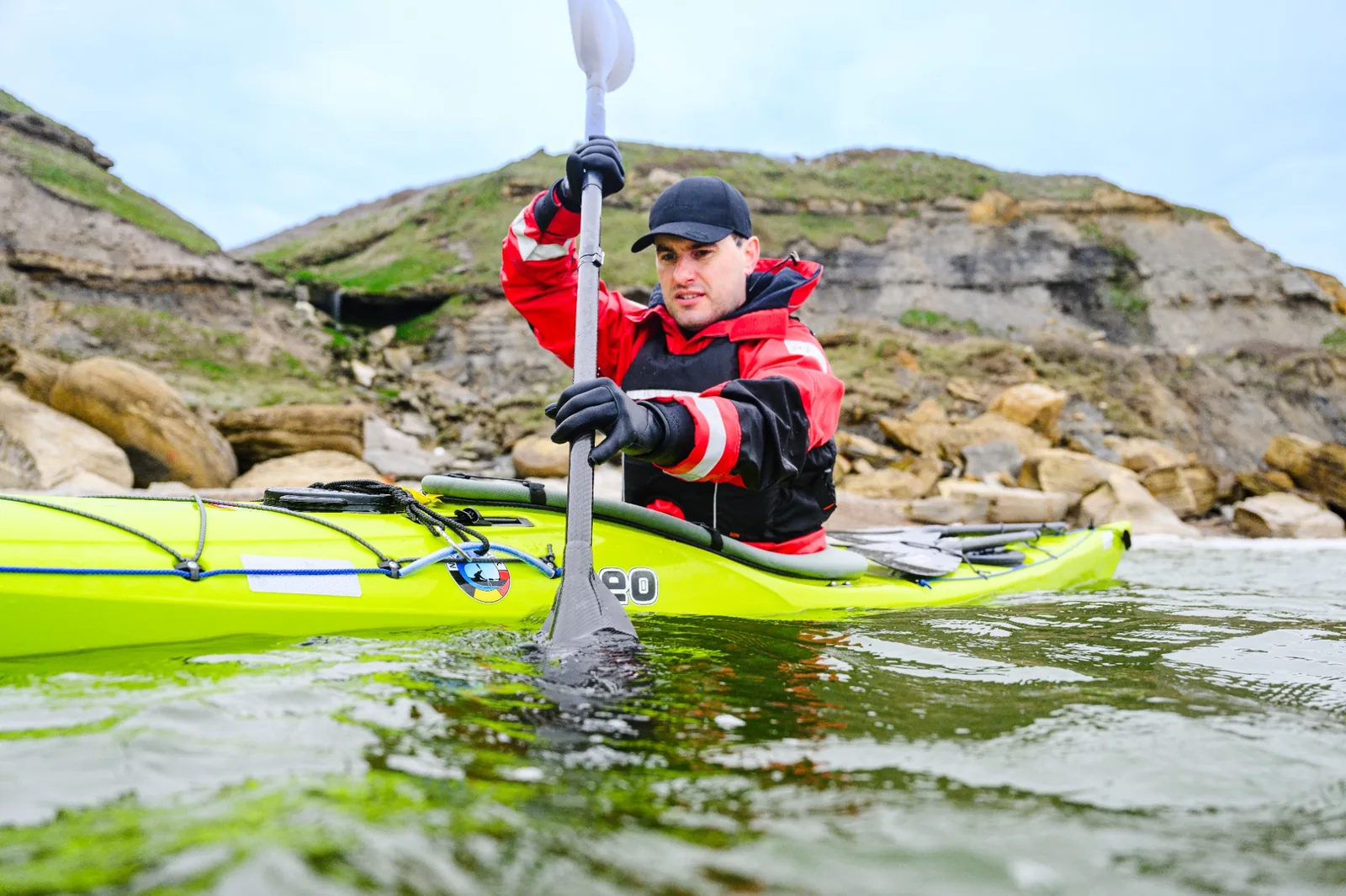 Sea kayaker in a red and black drysuit paddling a green touring kayak near a rocky shore
